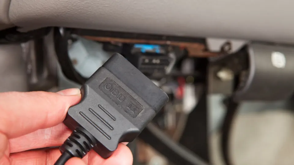Hand holding an OBD-II connector up to a car's diagnostic port under the dashboard